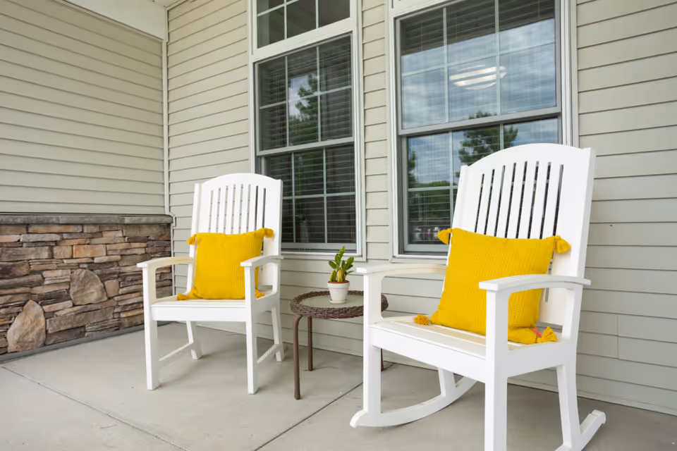 Two white rocking chairs with yellow pillows sit on a covered porch by windows with a small side table holding a potted plant.