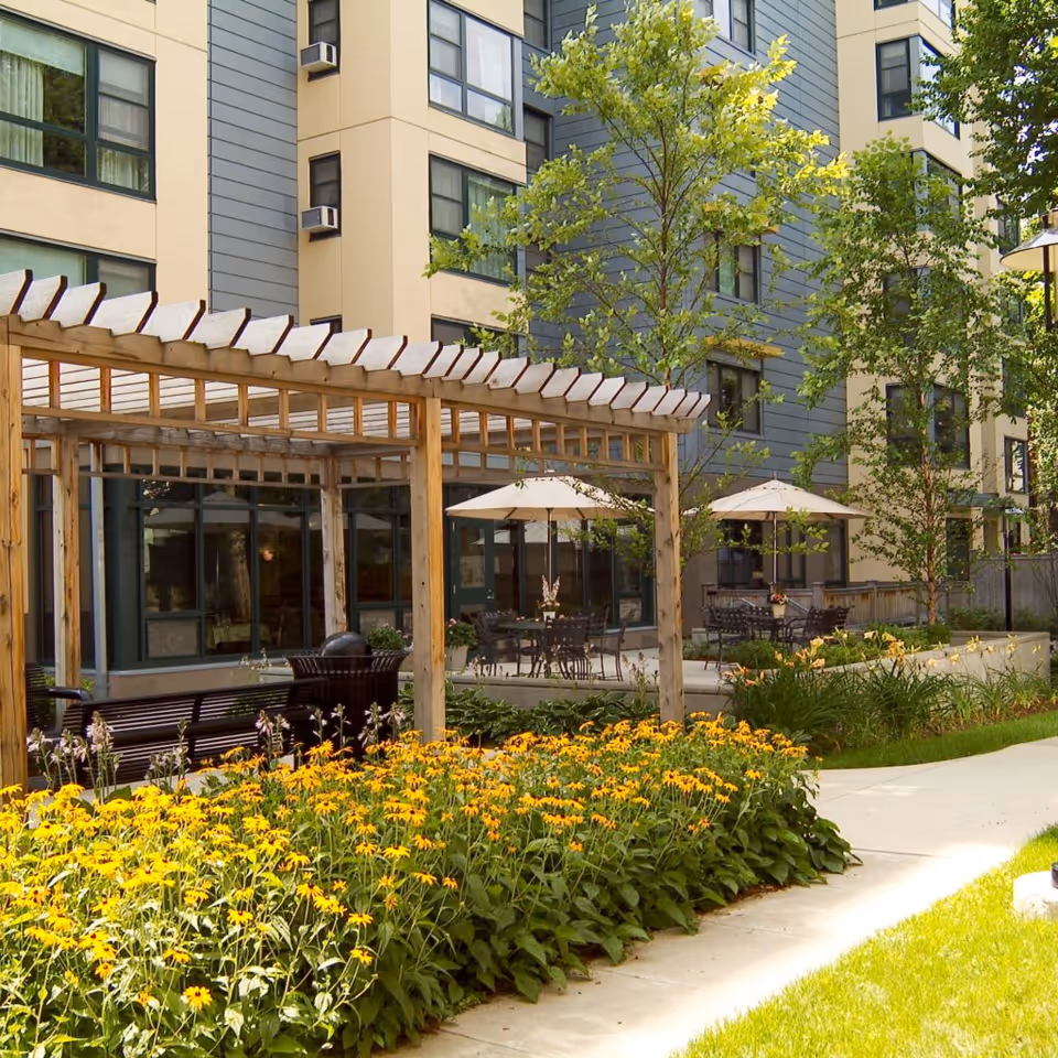 Outdoor garden area at JFK Apartments featuring a wooden pergola, yellow flowers, green trees, patio tables with umbrellas, and a modern multi-story building in the background.