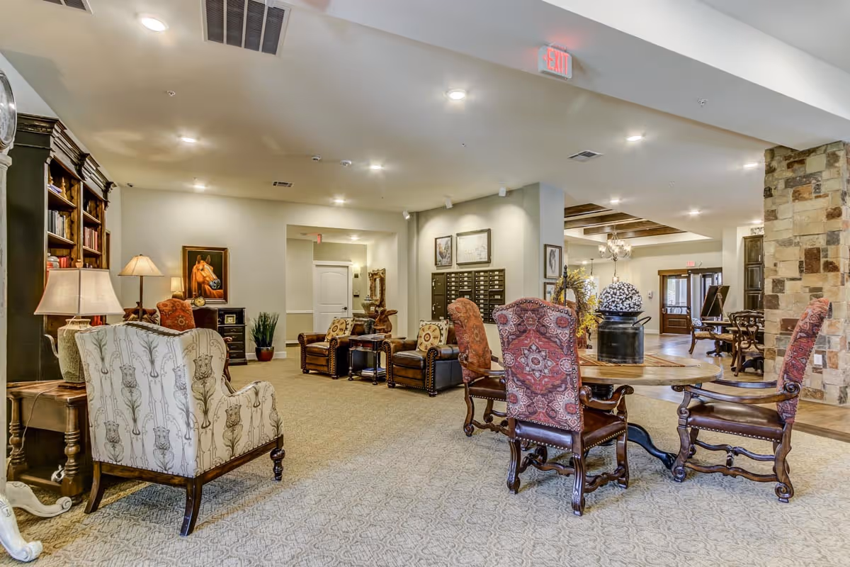 A spacious and well-lit living room area in an assisted living facility featuring a variety of upholstered chairs and armchairs arranged around wooden tables. The room has a carpeted floor, a stone pillar, bookshelves, lamps, framed artwork on the walls, and an exit sign visible on the ceiling.