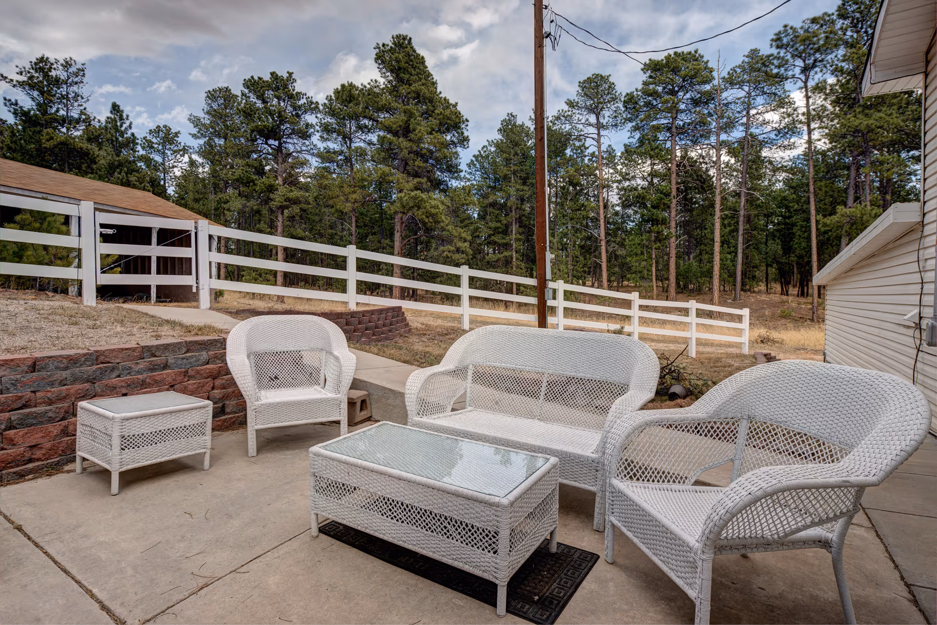 Outdoor patio area with white wicker furniture including two armchairs, a loveseat, a coffee table with glass top, and a side table. The patio is surrounded by a white wooden fence and trees in the background under a partly cloudy sky.