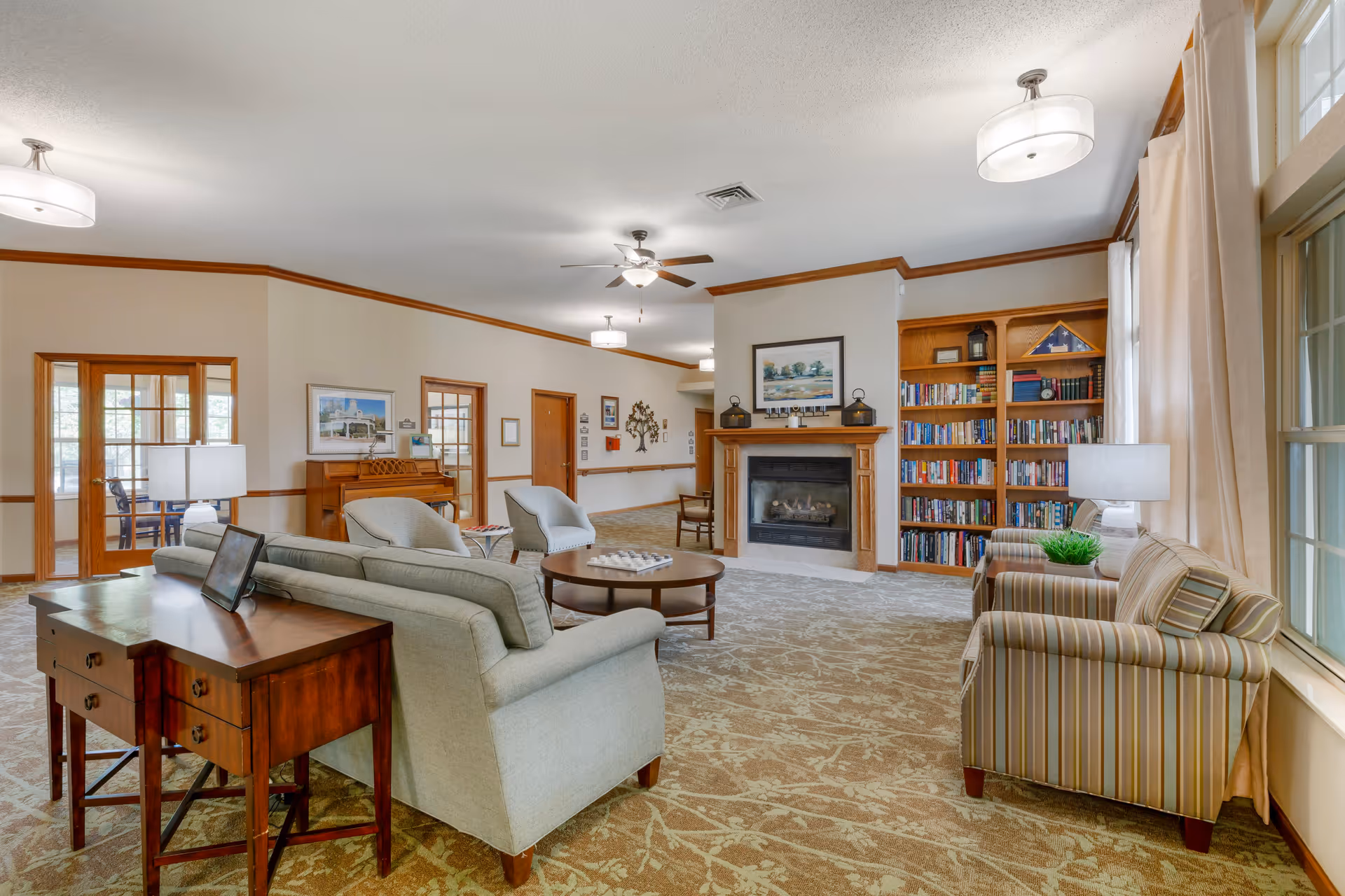A spacious and well-lit living room area in a senior living facility featuring comfortable seating including a light gray sofa, two armchairs, and a striped armchair near a window with beige curtains. The room has a carpeted floor with a floral pattern, a wooden coffee table, a fireplace with a framed picture above it, and a built-in bookshelf filled with books. There are ceiling lights and a ceiling fan, and a wooden piano is visible against one wall.