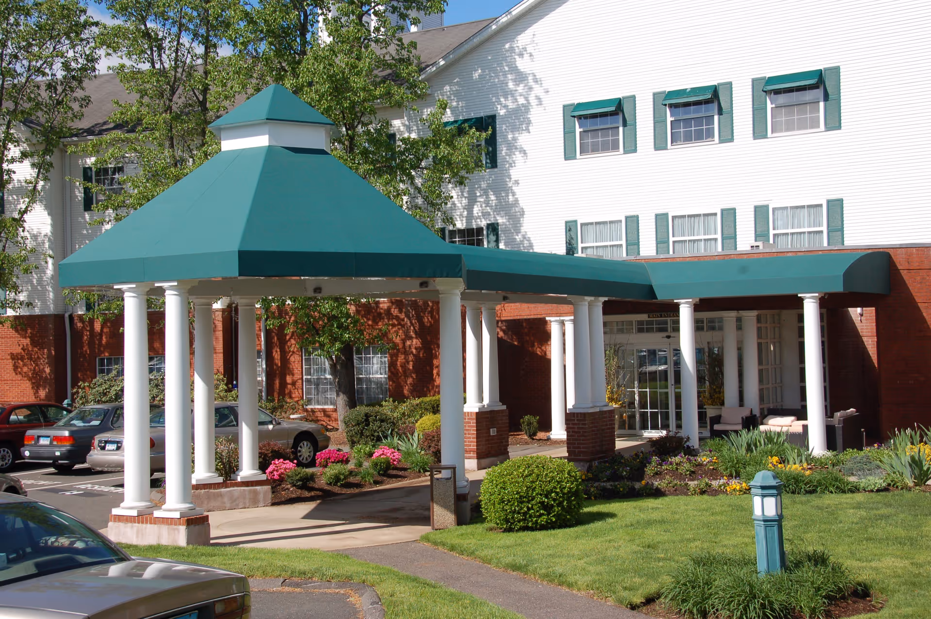 Entrance of a senior living facility with a green canopy supported by white columns, surrounded by landscaped bushes and flowers, with a multi-story building in the background featuring white siding and green window shutters.