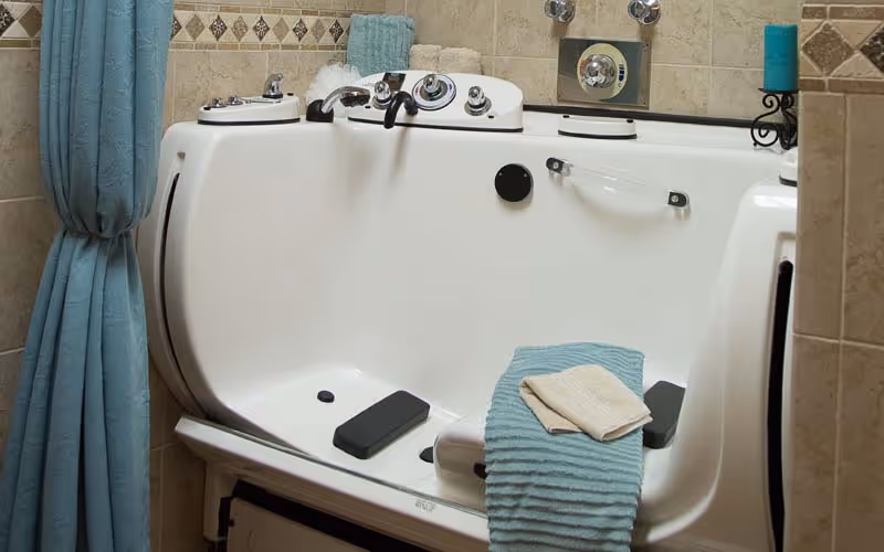 A walk-in bathtub with built-in controls and a seat, surrounded by beige tiled walls with decorative trim. A blue towel and a beige washcloth are draped over the edge of the tub, and a blue curtain is tied back on the left side.