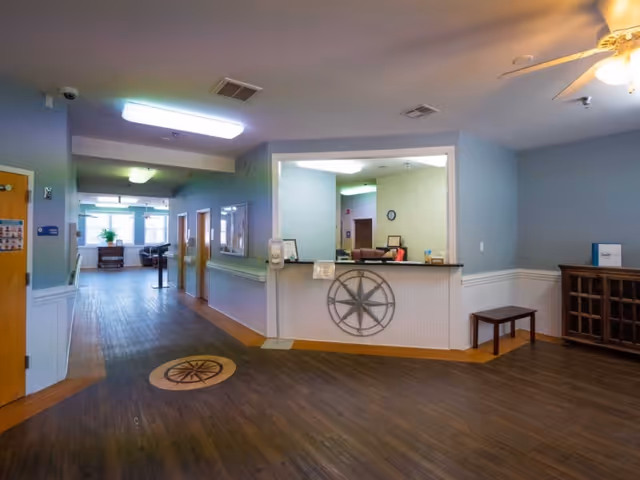 Reception area inside a senior living facility with a front desk decorated with a compass motif, a hallway leading to a seating area, wood floors and light blue walls.