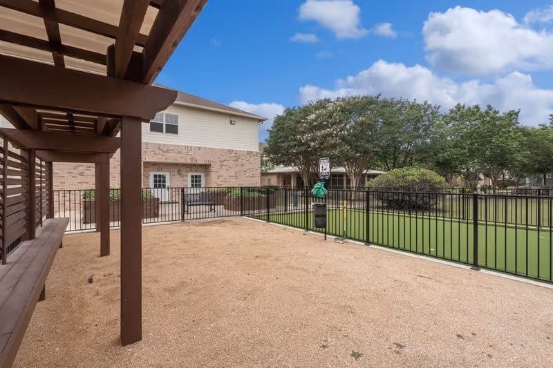 Outdoor fenced area with a gravel ground and a wooden pergola structure on the left side. Beyond the fence is a green artificial turf area, trees, and a two-story brick and siding building under a partly cloudy blue sky.