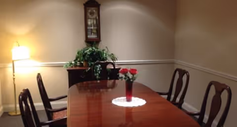 A dining room with a polished wooden table surrounded by six chairs. A vase with red flowers sits on a white doily in the center of the table. In the corner, there is a floor lamp providing warm light, a wooden cabinet with a green leafy plant on top, and a wall-mounted grandfather clock above the cabinet. The walls are beige with white trim.