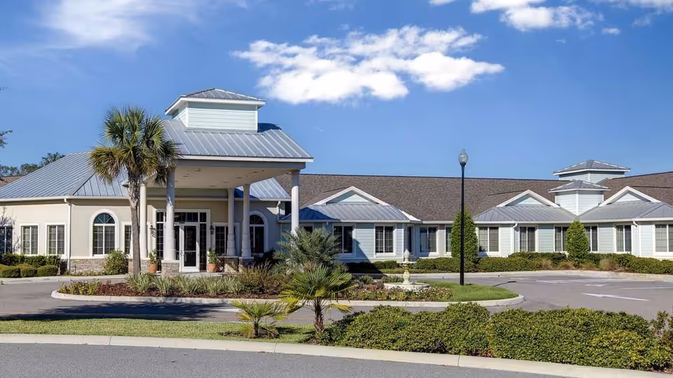 Exterior view of Lady Lake Senior Living facility showing a single-story building with a covered entrance, palm trees, landscaped bushes, and a clear blue sky with some clouds.