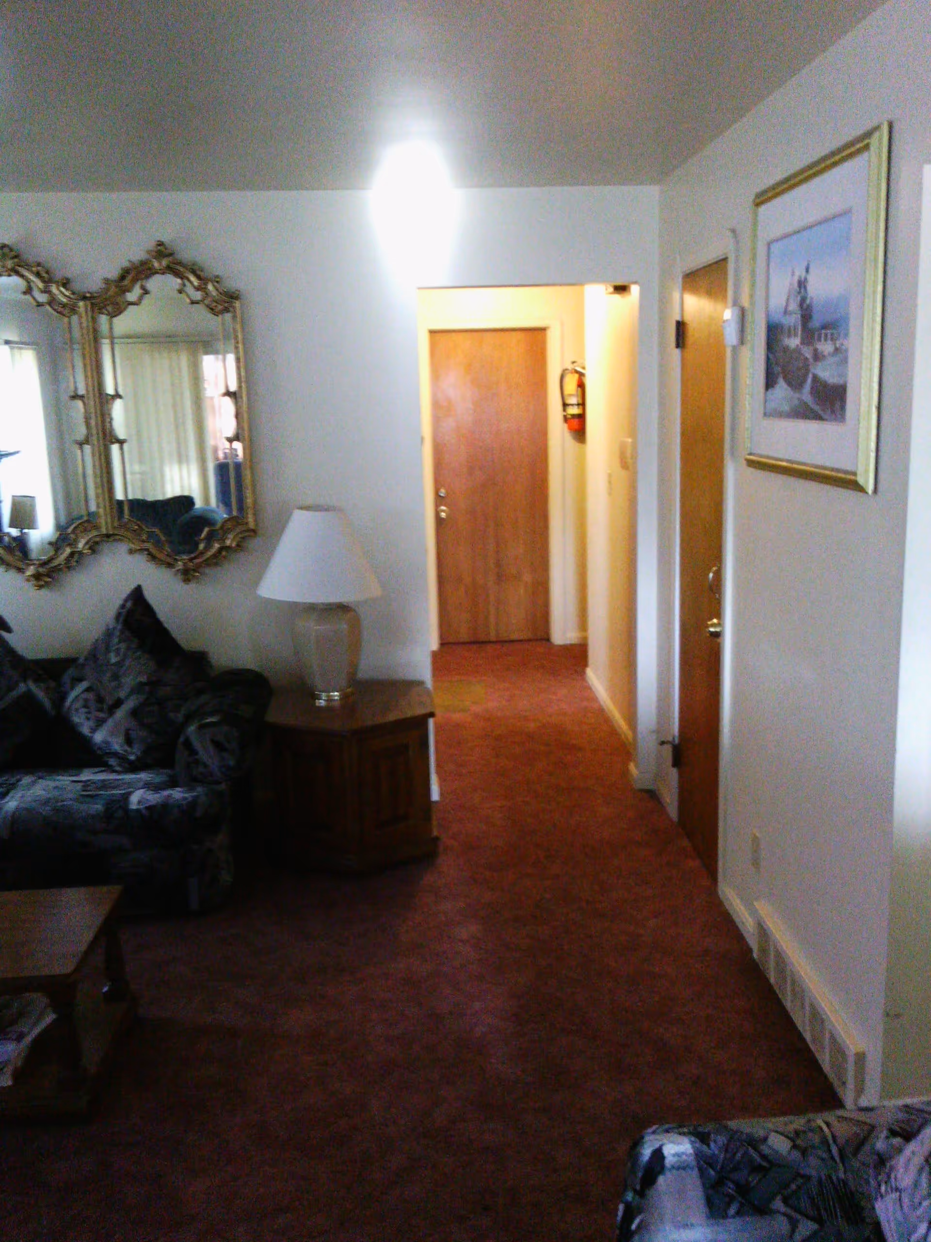 Interior view of a living room area with a patterned sofa, a wooden side table with a lamp, a large decorative mirror on the wall, and a hallway leading to a wooden door. The walls are white and there is a framed picture on the right wall.
