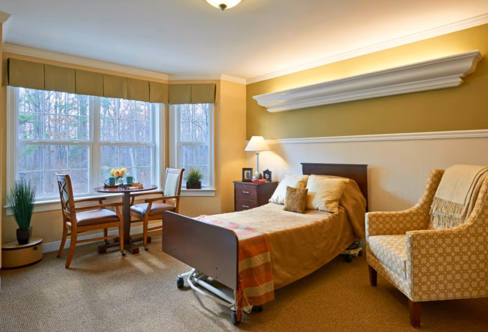 A cozy bedroom in a senior living facility with a single bed covered in beige bedding, a wooden nightstand with a lamp and picture frames, a patterned armchair with a throw blanket, and a small round table with two chairs near large windows showing a wooded outdoor view.