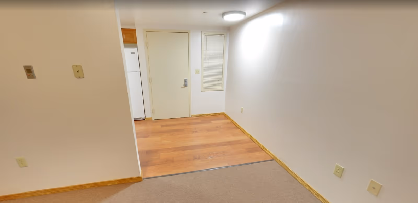 Interior view of a small room with beige carpet flooring transitioning to wood laminate flooring near a closed door and a small window with blinds. The walls are white with wooden baseboards, and a ceiling light fixture is visible. Part of a refrigerator and wooden cabinet can be seen through an opening on the left side.