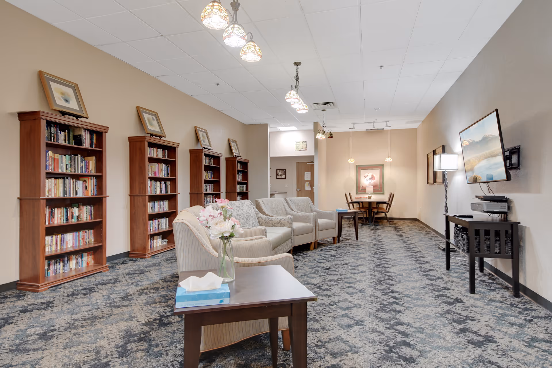 A spacious common area with a row of wooden bookshelves filled with books along the left wall. Several comfortable armchairs are arranged in the middle of the room with a small table holding a vase of flowers and a box of tissues. At the far end, there is a small dining table with four chairs under hanging pendant lights. On the right wall, a flat-screen TV is mounted above a console table with a lamp. The room has patterned carpet flooring and a white ceiling with multiple light fixtures.