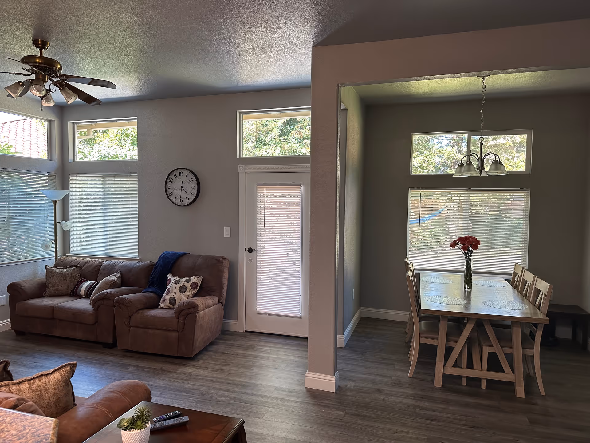 Open-plan living room and dining area with brown sofas, a wall clock, and a dining table topped with a vase of flowers.