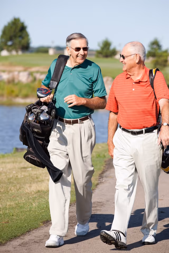 Two elderly men walking on a golf course path carrying golf bags, smiling and talking to each other on a sunny day with trees and water in the background.