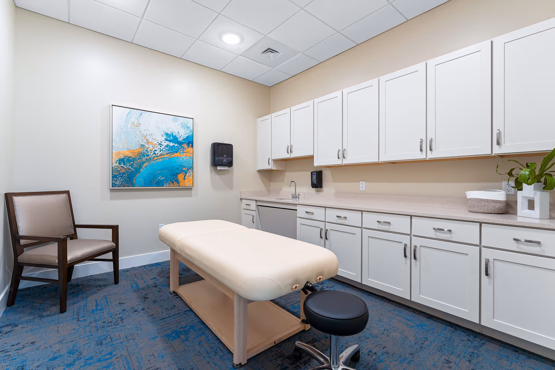 A clean and well-lit therapy or treatment room with a padded examination table in the center, a black rolling stool, a wooden chair with beige cushions, white cabinets along the wall, a countertop with a sink, a paper towel dispenser, a small plant, and a colorful abstract painting on the wall.