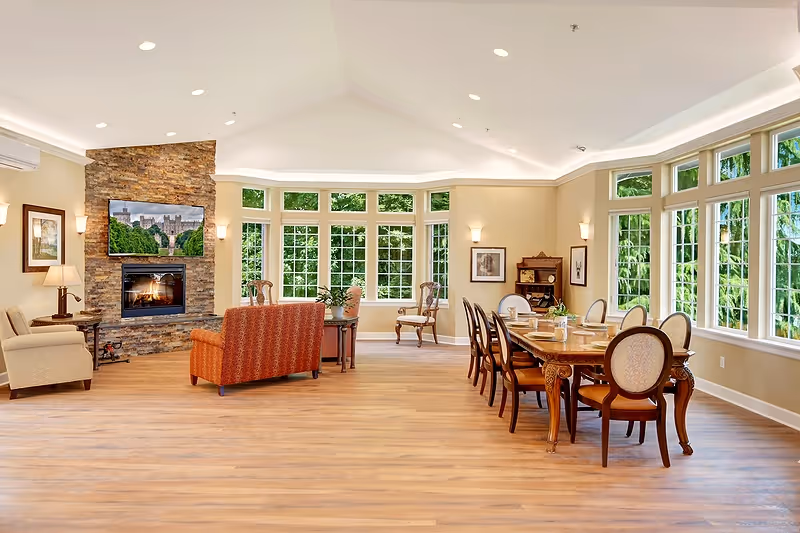 A spacious, well-lit room with large windows allowing natural light to fill the space. The room features a stone fireplace with a TV mounted above it, a seating area with armchairs and a sofa, and a long wooden dining table with chairs. The walls are painted a soft beige, and the floor is covered with light wood flooring. The room has a cozy and inviting atmosphere.