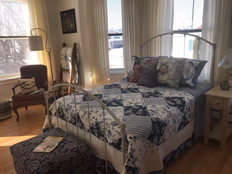 Sunlit bedroom with a metal-framed bed covered in a blue-and-white patchwork quilt, decorative pillows, a bedside table and a chair by the windows.