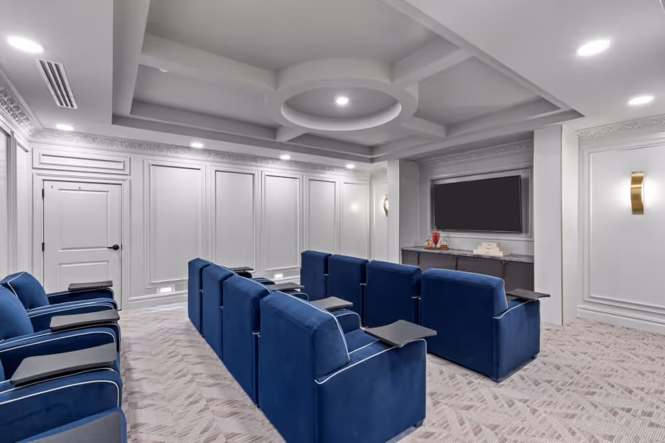 A small private theater room with two rows of blue upholstered recliner chairs, each equipped with a small black tray table. The room features white paneled walls, a coffered ceiling with recessed lighting, and a large flat-screen TV mounted on the front wall above a console table with decorative items.