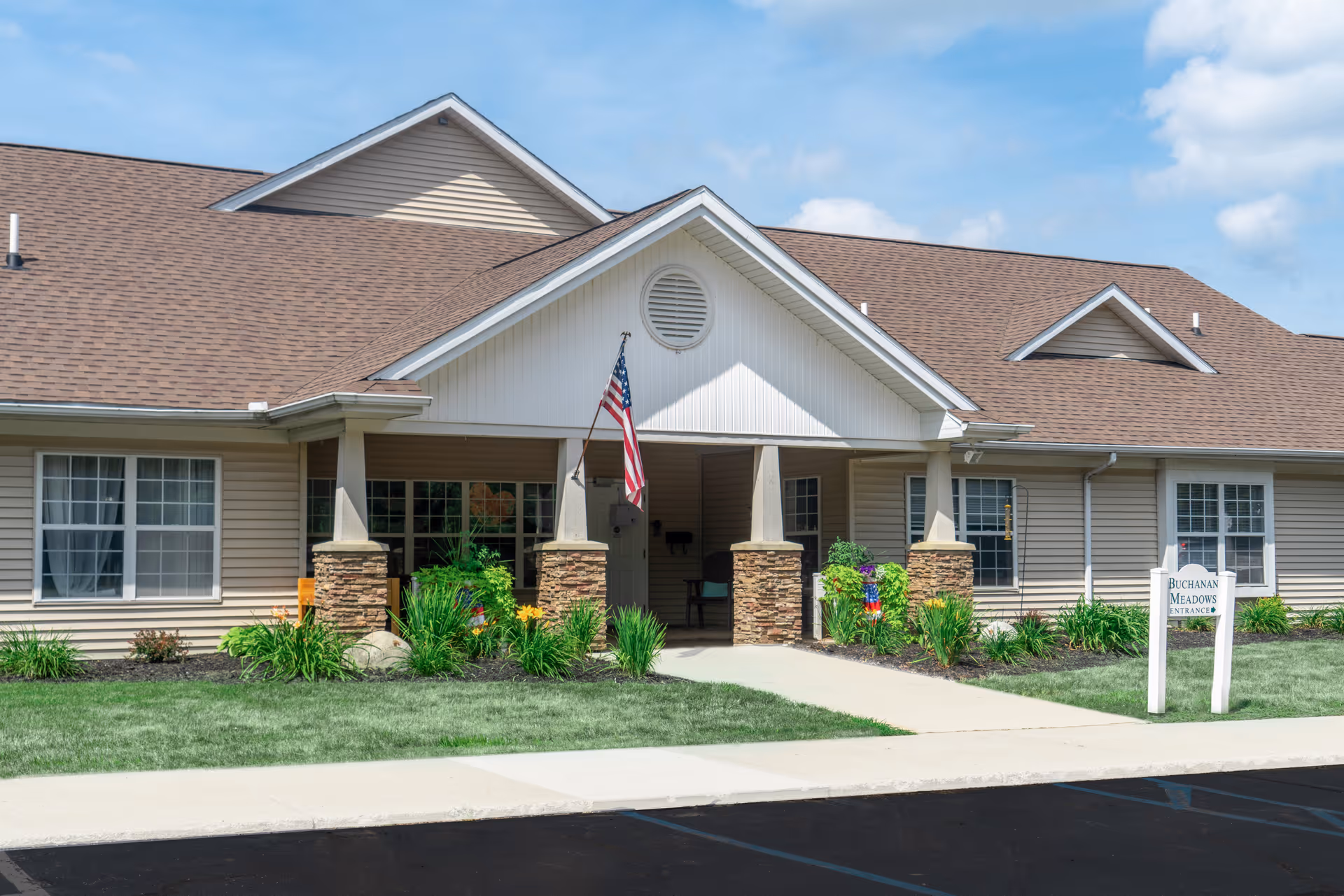 Front entrance of a single-story senior living building with a covered porch, American flag, landscaping, and a Buchanan Meadows sign.