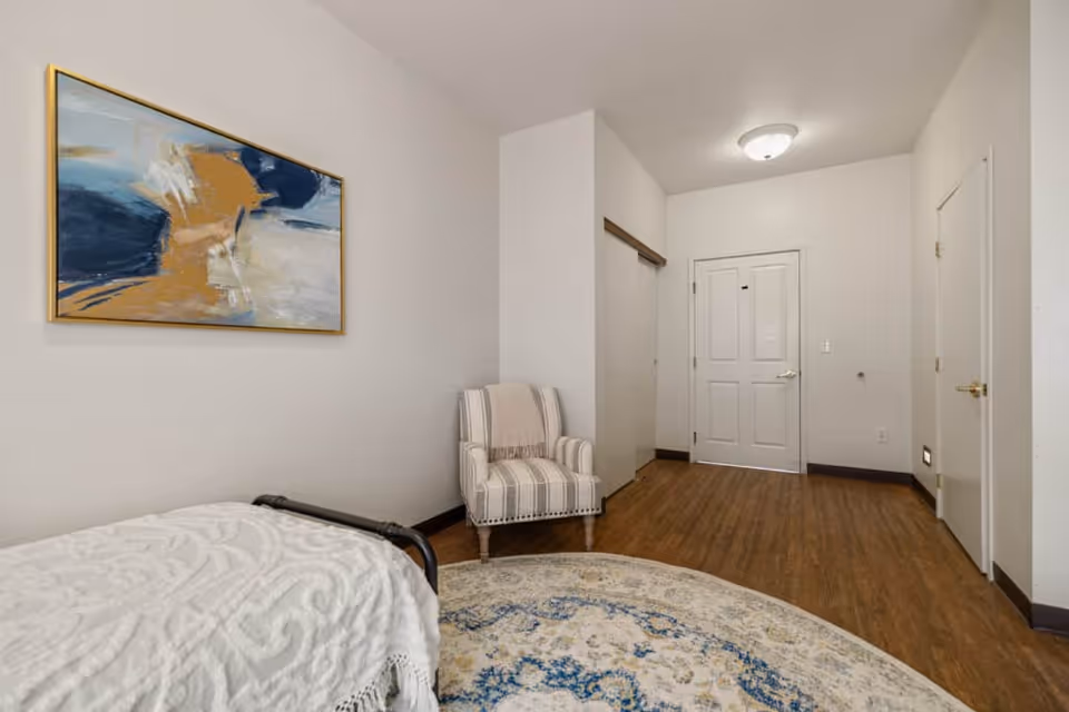 A simple, clean bedroom with a single bed covered in a white textured blanket, a striped armchair with a beige throw blanket, a round patterned rug on a wooden floor, and a large abstract painting with blue, white, and gold tones hanging on the wall. The room has white walls, a white door, and a closet with sliding doors.