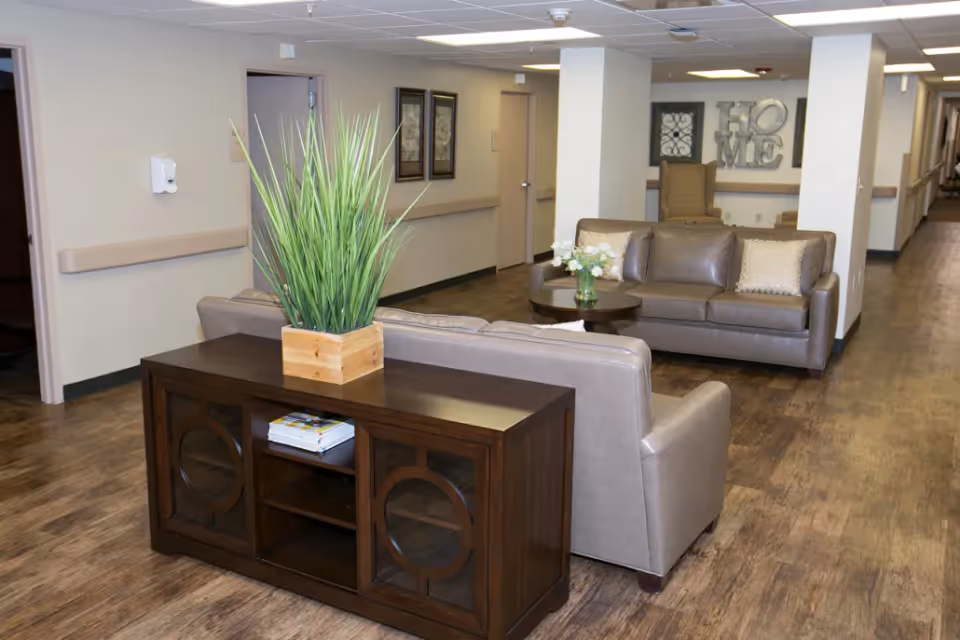 A cozy seating area in a senior living facility hallway with two beige leather sofas, a wooden console table with a potted plant, a round coffee table with a vase of white flowers, and wall decorations including framed pictures and a 'HOME' sign.