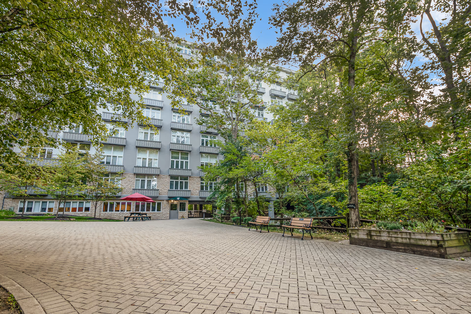 Paved courtyard with benches, trees, and a picnic table in front of a multi-story residential building.