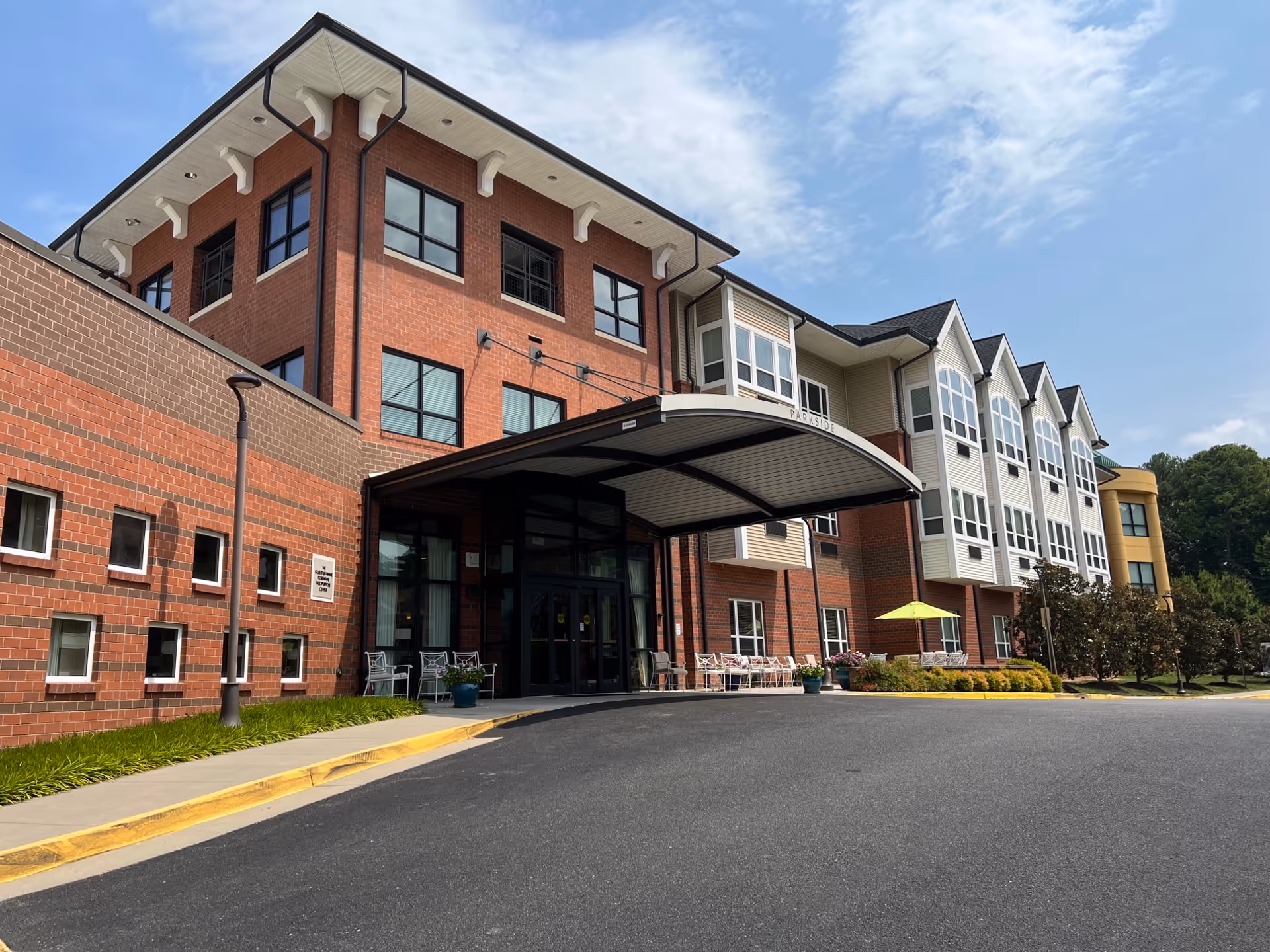 Exterior view of a multi-story senior assisted living facility building with a covered entrance, brick and siding facade, several windows, outdoor seating, and landscaping under a partly cloudy sky.