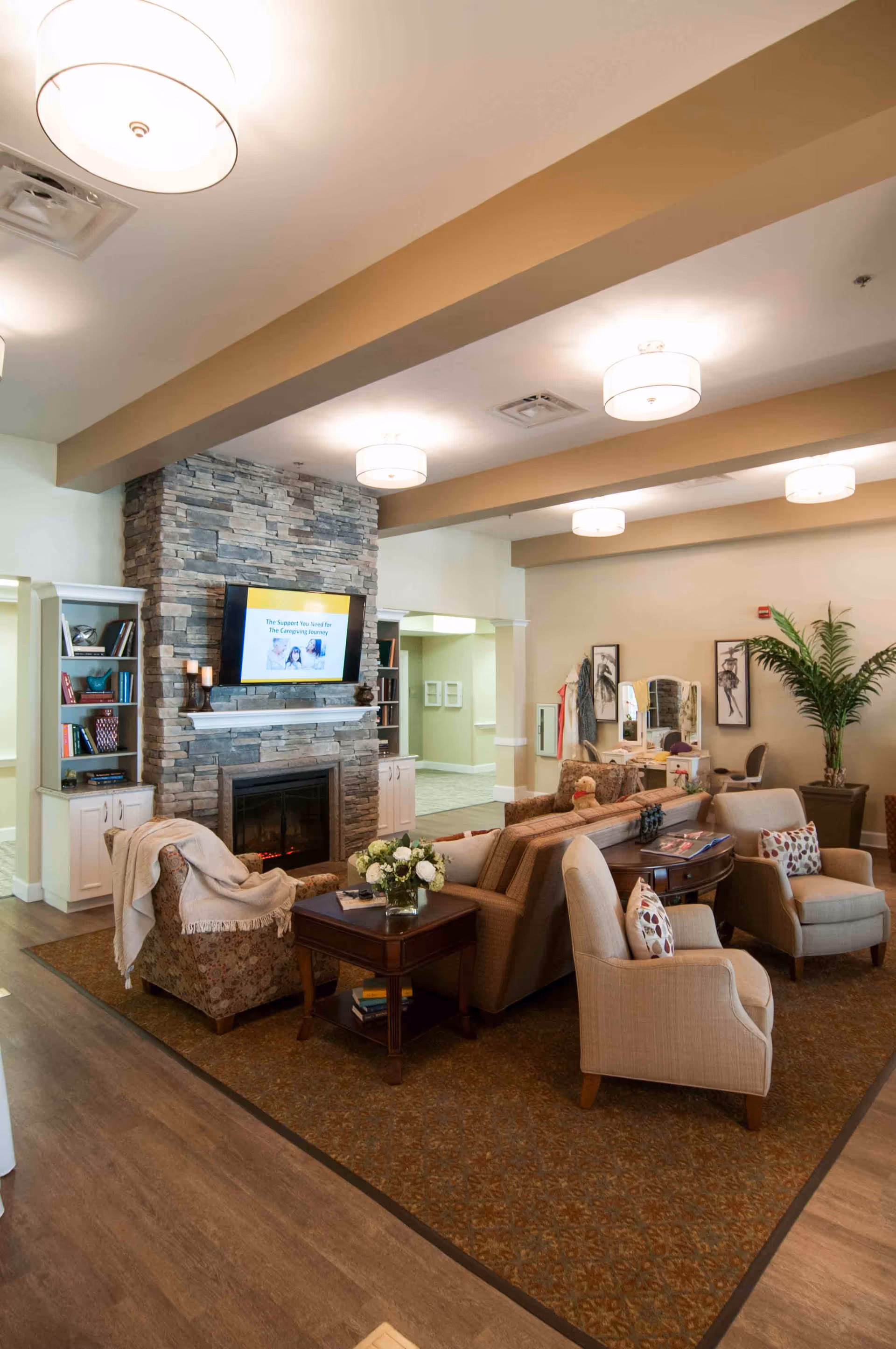 A cozy living room area with a stone fireplace and a mounted TV above it. The room features comfortable armchairs and a sofa arranged around a wooden coffee table with a vase of flowers. There are built-in shelves with books and decorative items on either side of the fireplace. The ceiling has exposed beams with modern light fixtures, and there is a large potted plant in the corner. The overall decor is warm and inviting.
