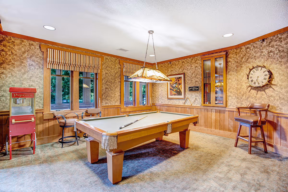 Wood-paneled game room with a pool table under a hanging light, seating, a popcorn machine and a wall clock.