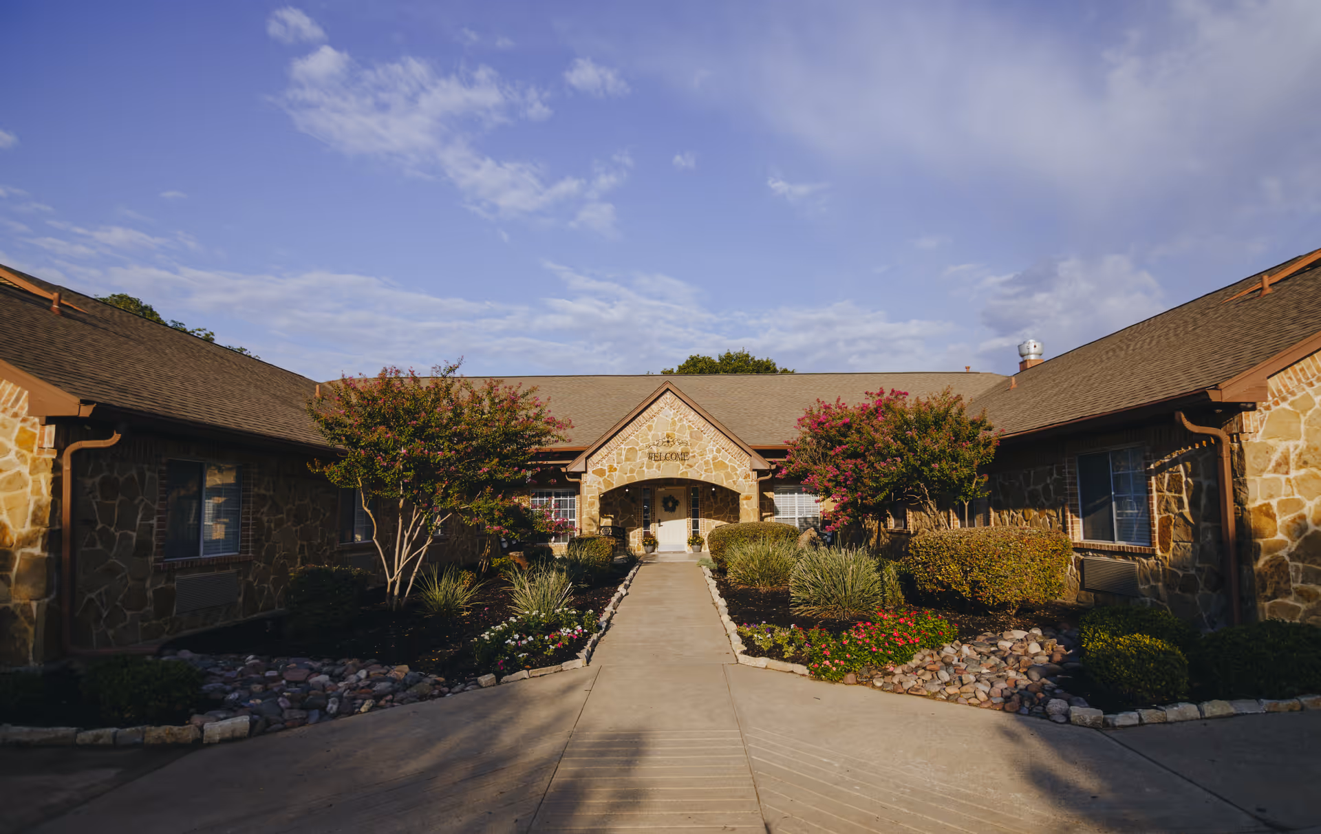 Front exterior view of a single-story stone building with a peaked roof and a central entrance. The entrance is framed by a small porch with a 'Welcome' sign above the door. There are landscaped flower beds with bushes and small trees on either side of a concrete walkway leading to the door. The sky is partly cloudy with blue patches.