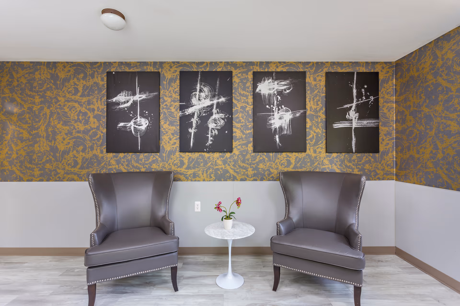 Two gray wingback chairs flanking a small round table with a potted plant beneath four abstract black-and-white paintings on patterned wallpaper.