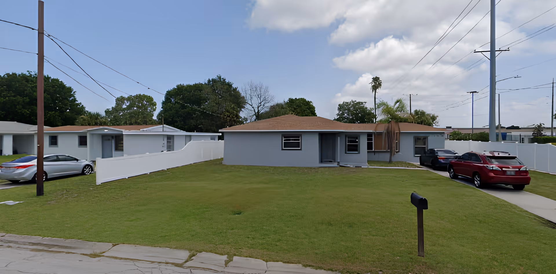 Single-story gray house with a large front lawn, driveway with parked cars and a mailbox under a partly cloudy sky.