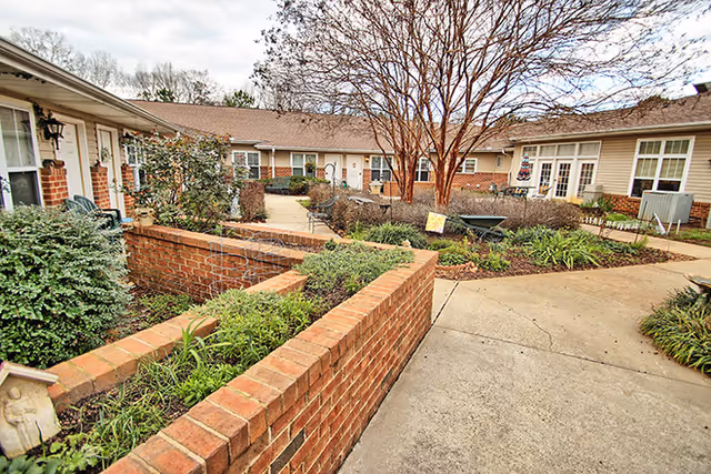 Outdoor courtyard area of a senior living facility with brick planters, shrubs, a leafless tree, and a paved walkway surrounded by single-story buildings with beige siding and white-framed windows.