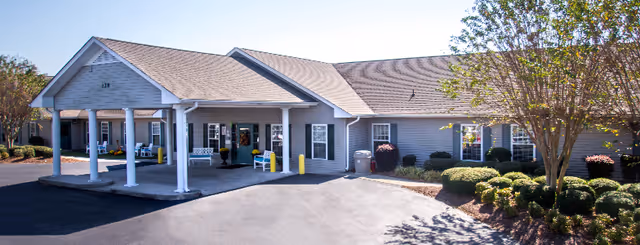 Front exterior of a single-story rehabilitation and nursing center with a covered entrance, driveway, benches, and landscaped shrubs.
