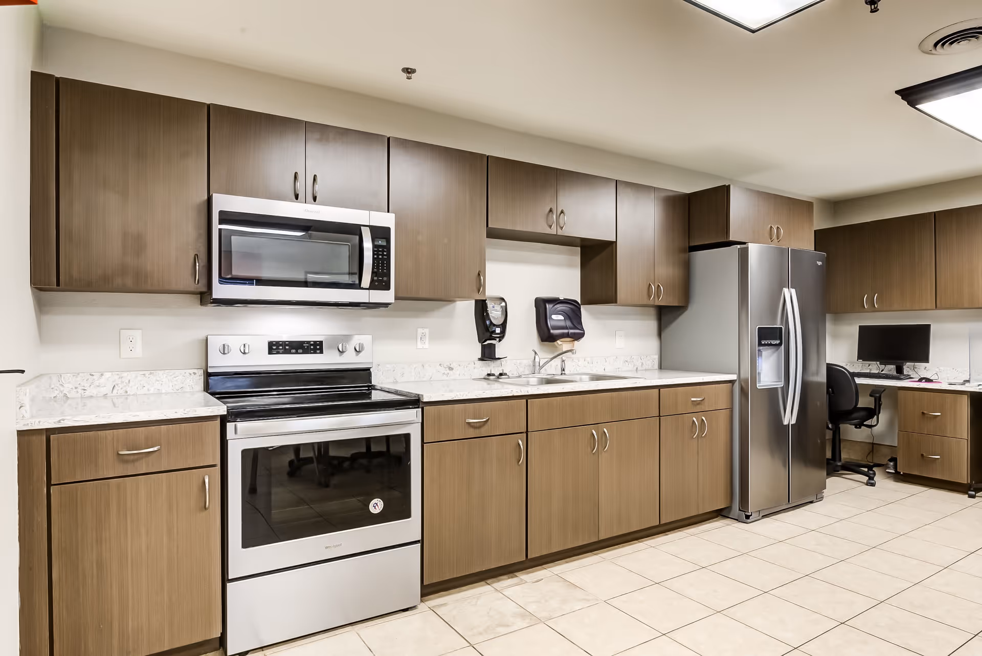A modern kitchen area with wooden cabinets, a stainless steel electric stove with oven, a microwave above it, a double sink with soap dispensers on the wall, and a stainless steel refrigerator. There is also a small office workspace with a computer and chair in the corner.
