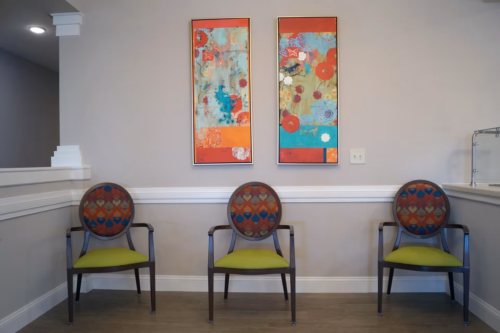 Three wooden chairs with colorful patterned backs and green seats are arranged against a light gray wall. Above the chairs, there are two vertical abstract paintings with red, orange, blue, and green tones. The floor is wooden, and there is a white chair rail molding on the wall.