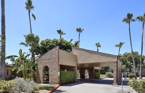 Exterior view of a nursing and rehabilitation center building entrance with a covered driveway, surrounded by palm trees and greenery under a clear blue sky.