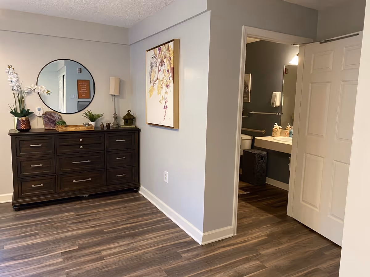 Interior view of a room with wood flooring and light gray walls. A dark wooden dresser with multiple drawers is against one wall, decorated with a round mirror, a lamp, plants, and small decorative items. A framed picture of an owl on a branch hangs on the adjacent wall. An open door reveals a bathroom with a sink, mirror, and toilet.