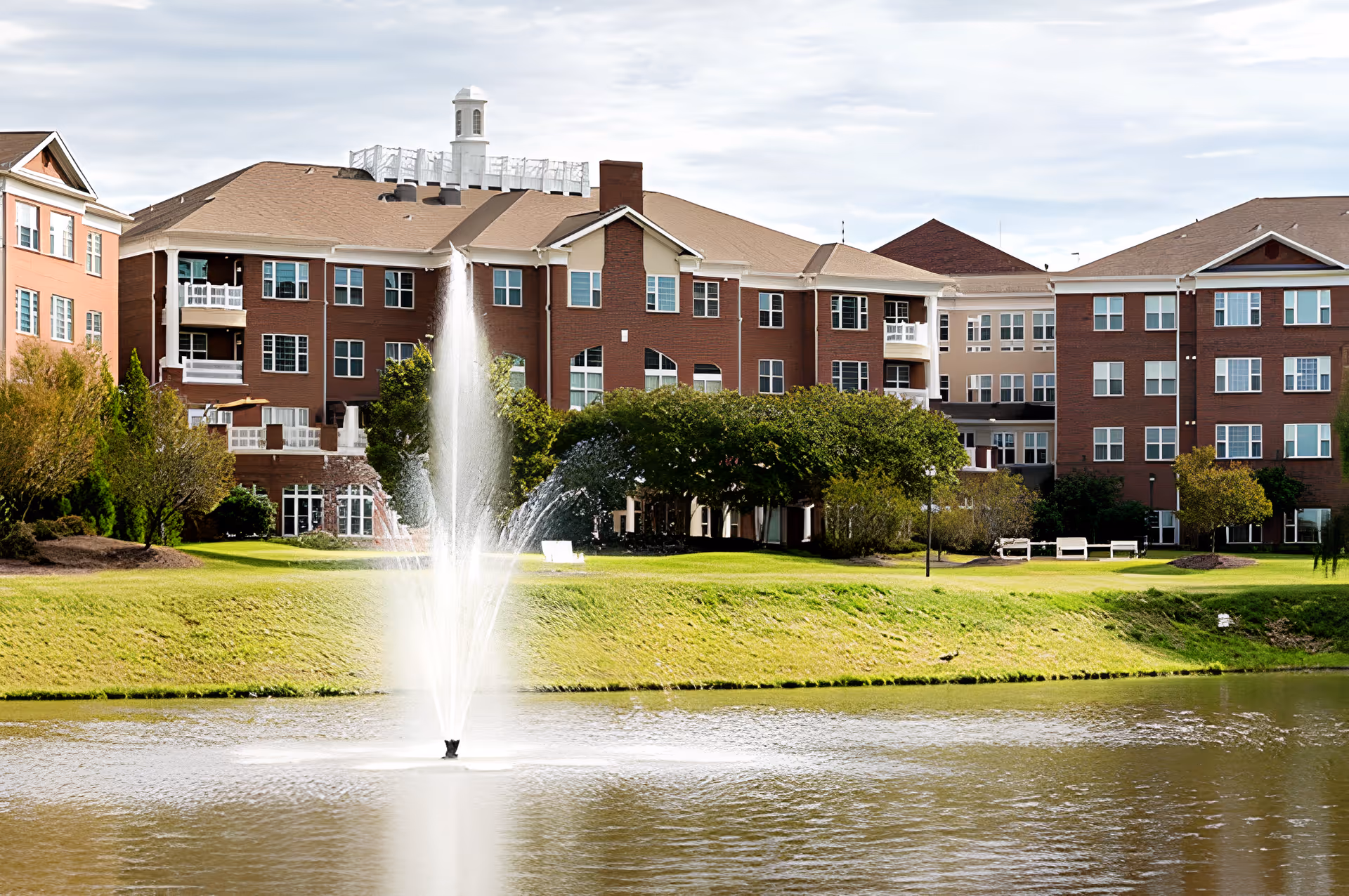 Brick senior living building seen from across a pond with a central fountain, lawns, trees, and benches.