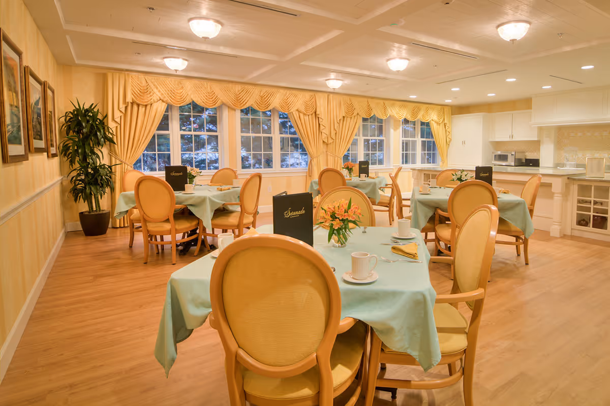 Bright dining room with round tables set with pastel tablecloths and chairs, large windows with yellow drapes and a serving counter in the back.