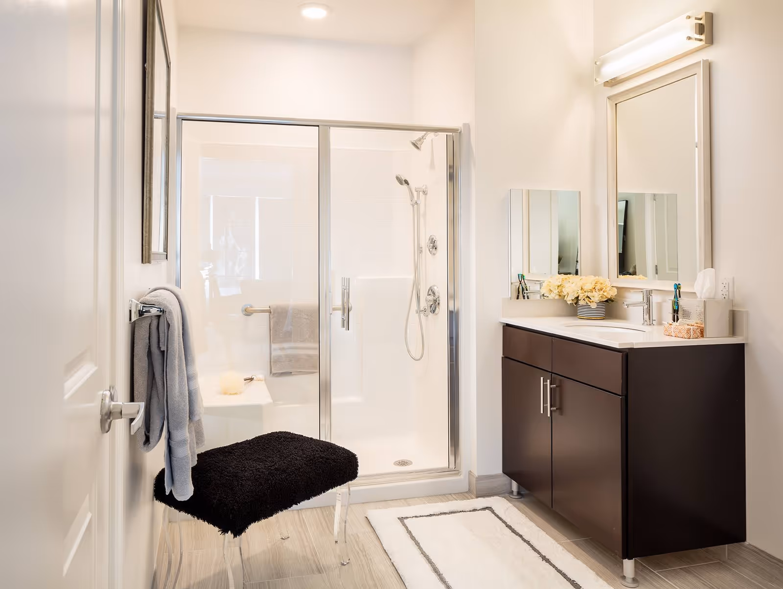 Bright bathroom with a glass-enclosed shower, a dark wood vanity with a white countertop, a mirror with lights above it, a black cushioned stool, and a towel hanging on the door.