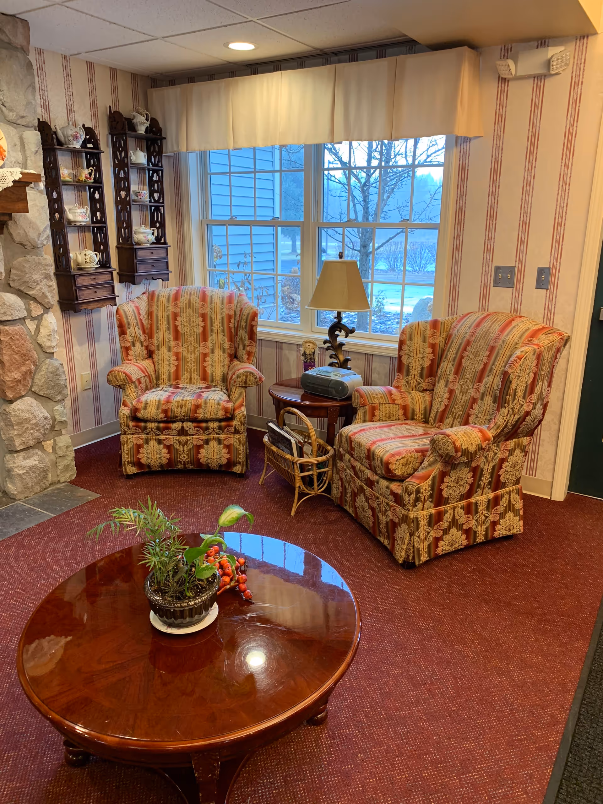 A cozy sitting area in a living room with two patterned armchairs, a wooden coffee table with a small potted plant and decorative berries, a side table with a lamp and a radio, and a window with a cream-colored valance. The walls have striped wallpaper and there is a stone fireplace partially visible on the left.