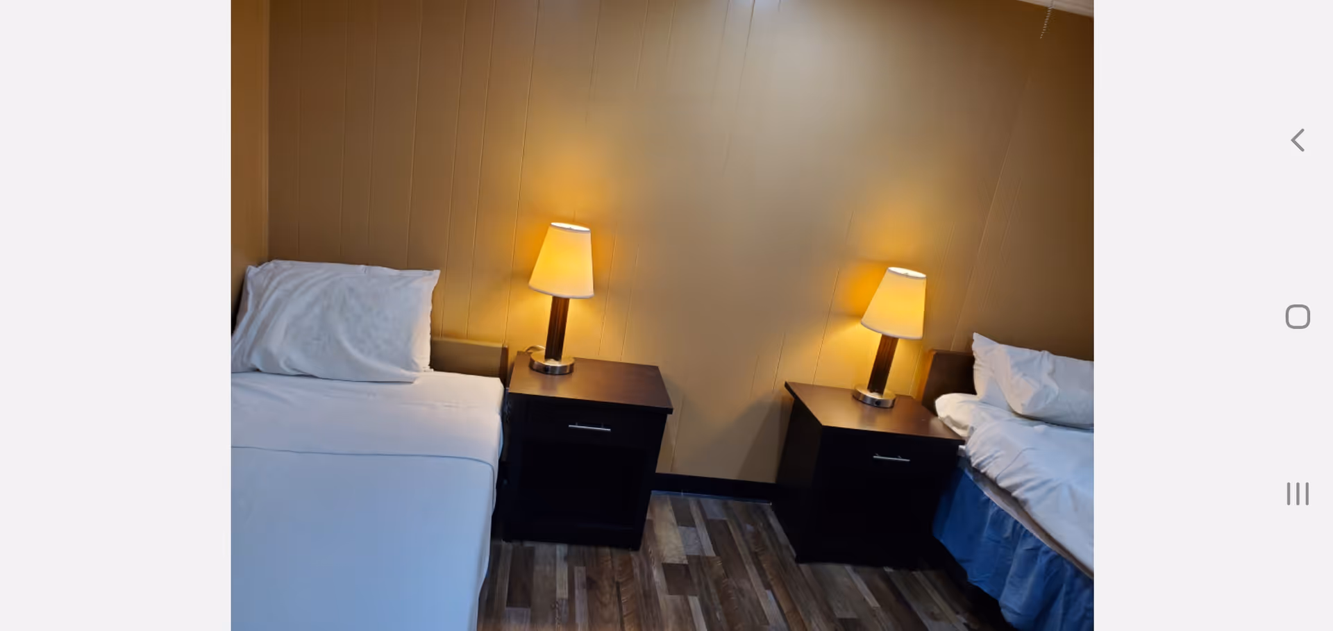 A room with two single beds separated by two dark wooden nightstands, each with a lit table lamp. The beds have white bedding and pillows, and the walls are painted a light brown color. The floor has a wood-like pattern.