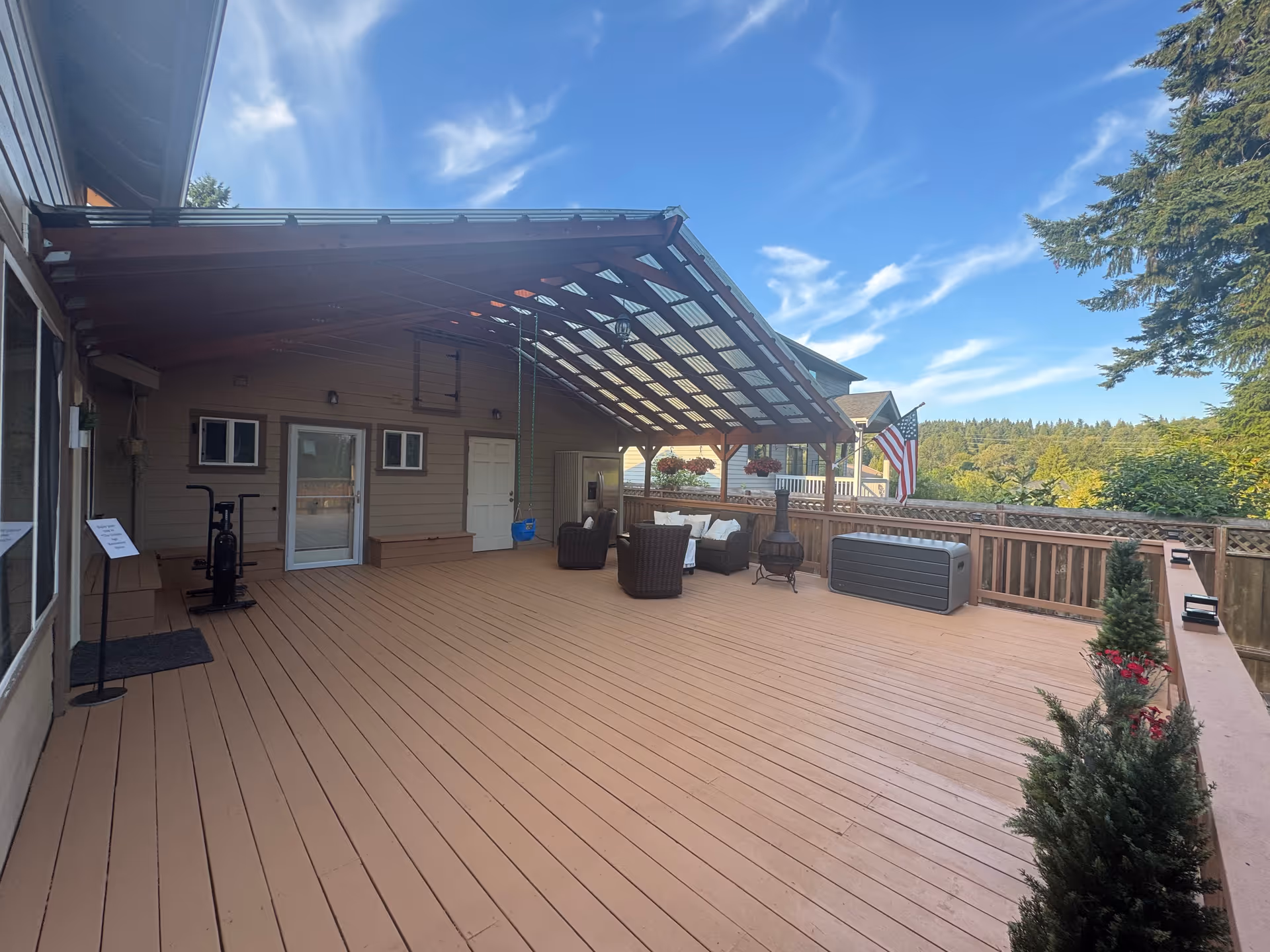 A spacious outdoor wooden deck attached to a building, featuring a covered seating area with wicker chairs and a small table. The deck has a swing, a chiminea, an American flag, and potted plants along the railing. The sky is clear with some clouds, and trees are visible in the background.