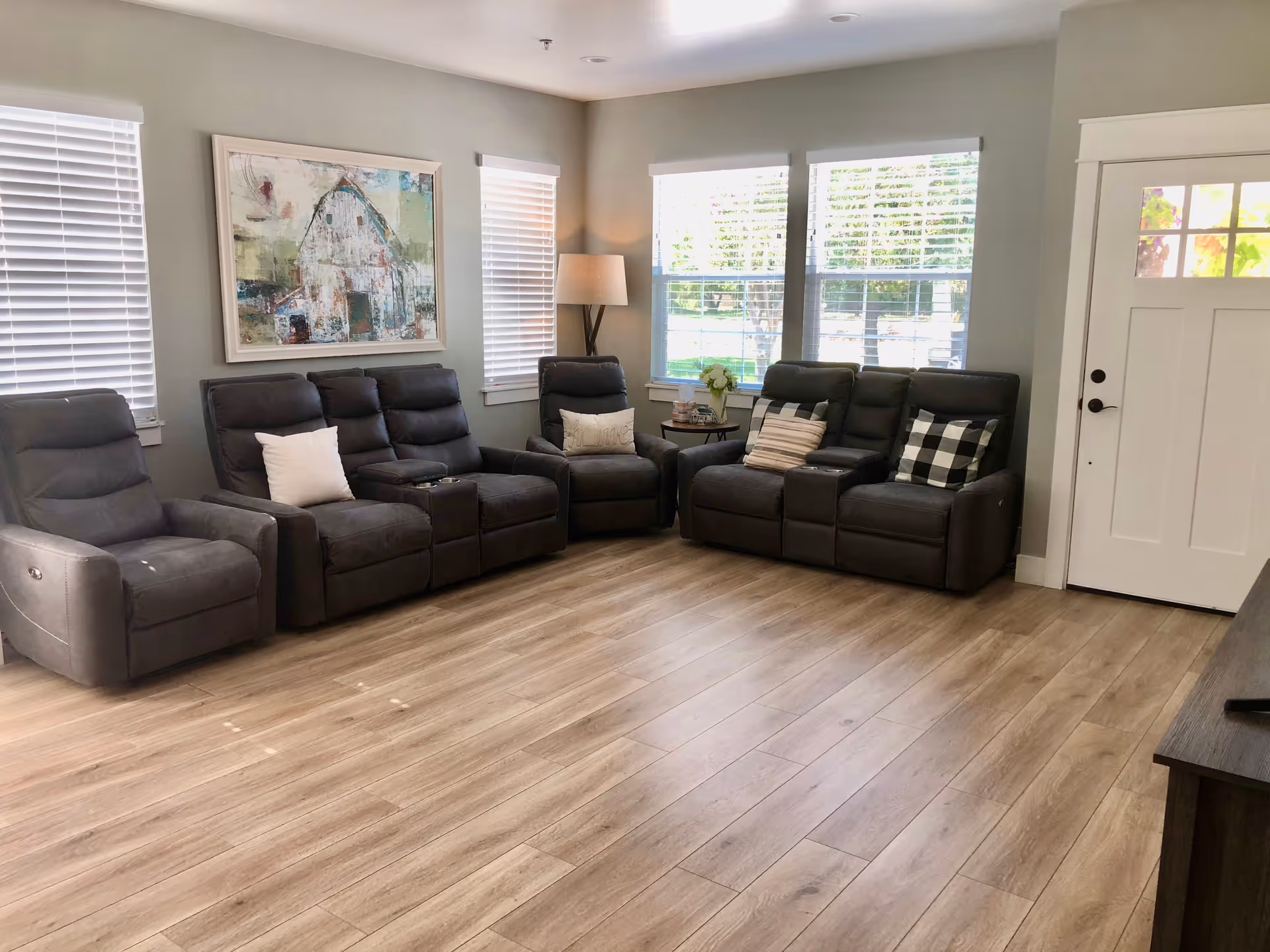 Spacious living room with multiple dark gray recliners, wood flooring, windows, and a white entry door.