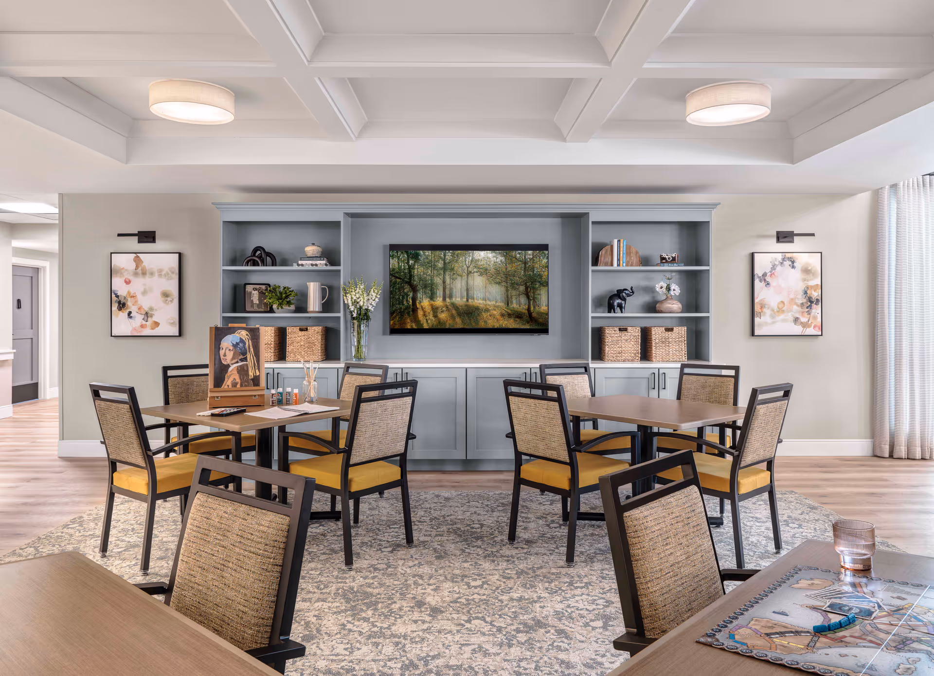 A bright and inviting common area with several tables and chairs arranged on a patterned rug. The back wall features built-in shelves and cabinets painted in light blue, decorated with books, baskets, plants, and art pieces. A large framed picture of a forest scene hangs in the center. The ceiling has a coffered design with two round light fixtures, and there are two abstract paintings on the side walls. A board game and a glass of water are visible on the table in the foreground.