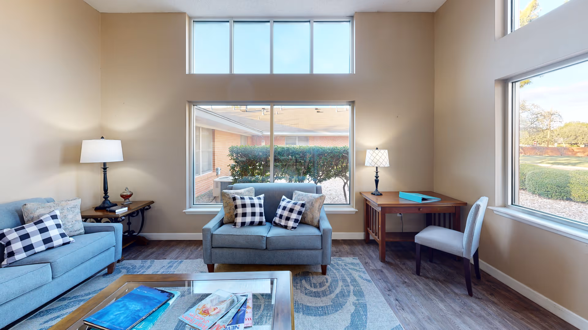 A bright living room with two blue sofas adorned with checkered and patterned pillows, a wooden coffee table with magazines, a side table with a lamp and books, and a wooden desk with a chair and a lamp. Large windows provide natural light and a view of greenery outside.