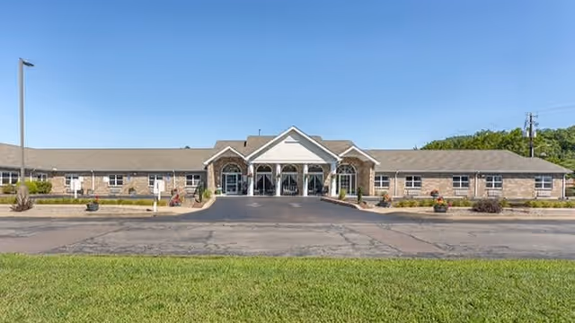 Front exterior view of a single-story senior living facility building with a covered entrance, multiple windows, and a paved driveway. The building is surrounded by a grassy area and some landscaping with shrubs and flowers under a clear blue sky.