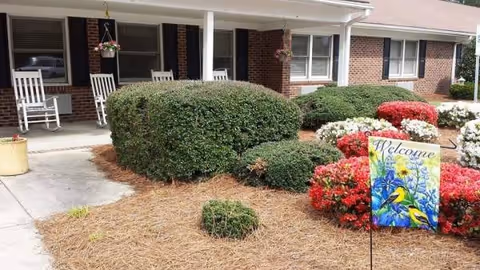 Brick senior living facility exterior with trimmed hedges, colorful flowers, and a welcome sign.