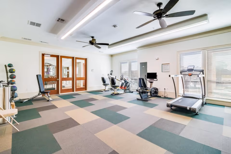 Bright exercise room with patterned carpet flooring, two ceiling fans, large windows with blinds, and various fitness equipment including a treadmill, stationary bikes, a weight bench, and a rack of medicine balls.