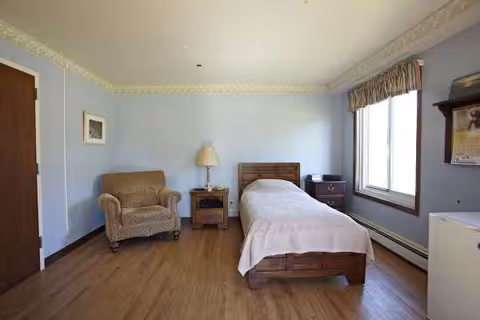 A single bedroom with a wooden bed, nightstands, chair, lamp, and window against blue walls.
